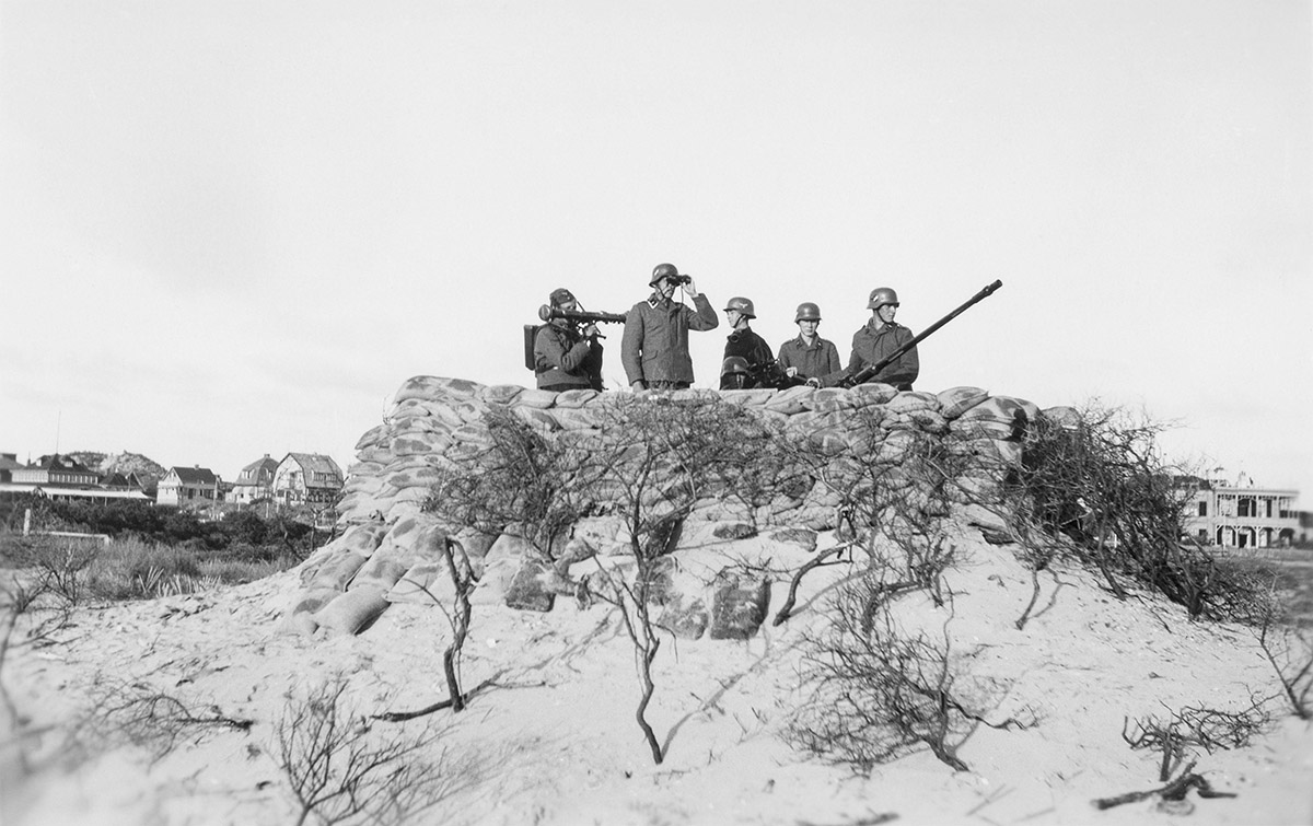 Luchtdoelgeschut langs de Nieuwe Waterweg, Hoek van Holland, juni 1940<br>
Uit het album van soldaat Salthammer / collectie Peter de Krom<br>
Walter Salthammer<br>
Gefreiter Reserve Flakabteilung 115<br>
Amateurfotografie werd in het Duitse leger aangemoedigd als ontspanning voor de soldaten. Zo fotografeerde soldaat Walther Salthammer in juni 1940 zijn eenheid luchtdoelgeschut in de duinen voor de Oude Hoek.<br>
Het is een unieke foto, omdat het oude dorp binnen een kilometer van het strand lag, in verboden militair gebied. Om die reden werd het vanaf 1942 gesloopt. Rechts staat een van de eerste gebouwen van het dorp, het kantoor van Rijkswaterstaat. Het privé-album van Salthammer geeft een optimistisch en ontspannen beeld van het dagelijks leven van de soldaten met veel soldatenhumor, sport, meisjes en bier. In 1941 vertrok hij met zijn eenheid naar Frankrijk.