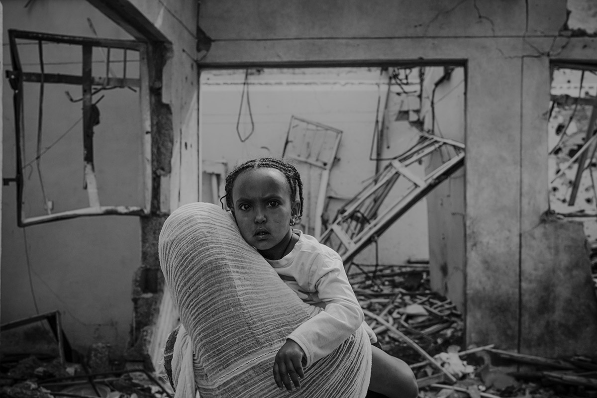 Heran Alemayo, Tigray, Ethiopië<br>
Lieuwe Siebe de Jong - 2023<br>
Heran ligt op de schouder van haar moeder, Birhan Hagos, terwijl ze door hun verwoeste huis lopen. Het werd getroffen tijdens een aanval op de avond dat het officiële vredesakkoord tussen de regering van Ethiopië en het Tigray People's Liberation Front (TPLF) werd ondertekend. In de dagen die volgden, bleef de situatie gespannen. De oorlogsschade is enorm. Herans vader werkte hard om zijn huis te kunnen betalen; net vóór de oorlog was het klaar. Nu heeft het gezin geen huis meer om naar terug te keren.