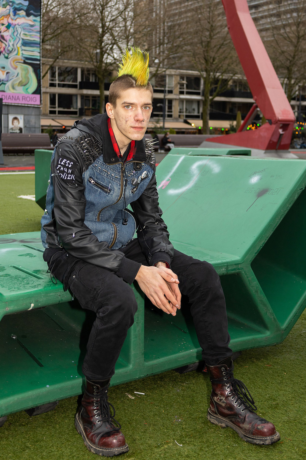 Petra Boegheim (Diemen), 1e prijs, Samenleven doe je zo?<br>
'Deze jongeren die ik trof op straat in Rotterdam. Allemaal hun eigen onderscheidende stijl. Waren zij er bij toeval of verbleven zij daar omdat er geen andere, vrije plek was tijdens deze zoveelste lockdown?<br>
Een jonge punker, zittend bij de skatebaan in Rotterdam centrum. Zijn felgekleurde hanenkam staat stoer recht op zijn hoofd. Een verlegen blik, afstandelijk maar tegelijkertijd nieuwsgierig. Zijn armen in gesloten houding, afwachtend op wat komen gaat. Het lijkt alsof deze jongen uit een andere wereld komt. Het beeld is een subtiel commentaar op het thema van de inclusieve samenleving waarin iedereen zichzelf wil en mag zijn.'